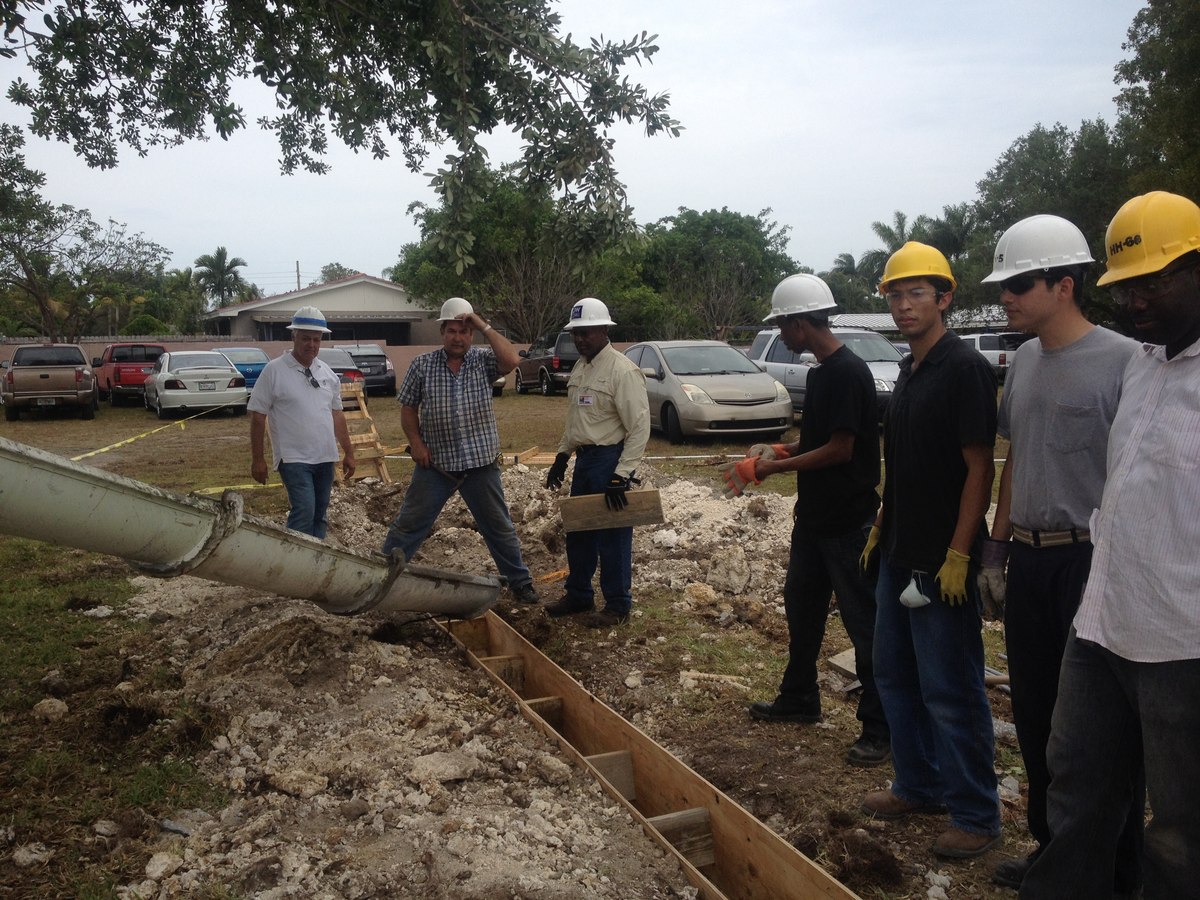 Concrete chute pouring into a wood-formed footer trench with eight crew in white and yellow hard hats coordinating the pour.