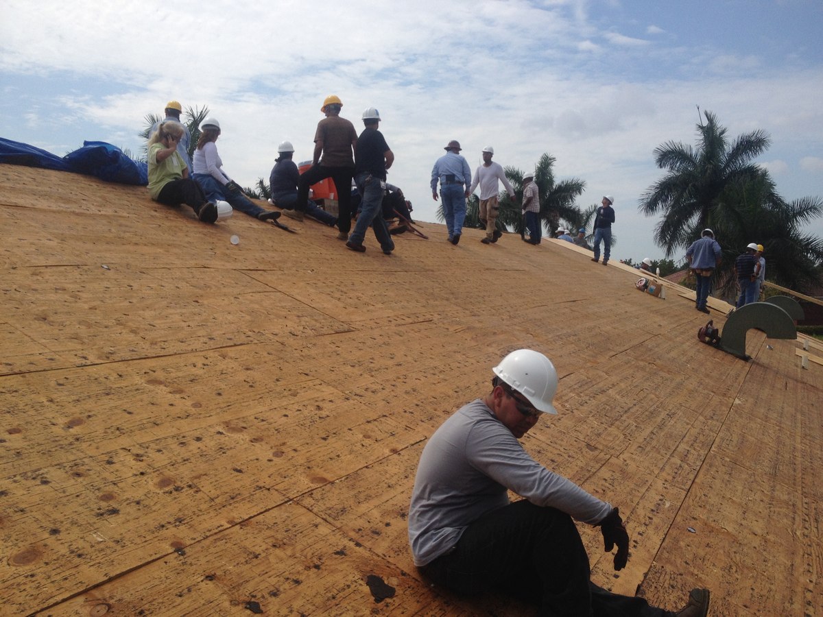 View along a plywood-sheathed roof deck with twelve workers in hard hats spread across a residential reframe in South Florida.