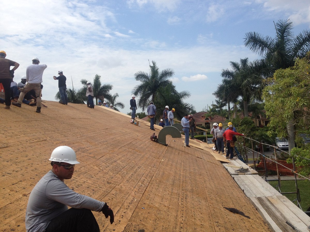 Foreman in white hard hat in foreground, twelve workers spread across a plywood-sheathed residential roof reframe under blue sky.