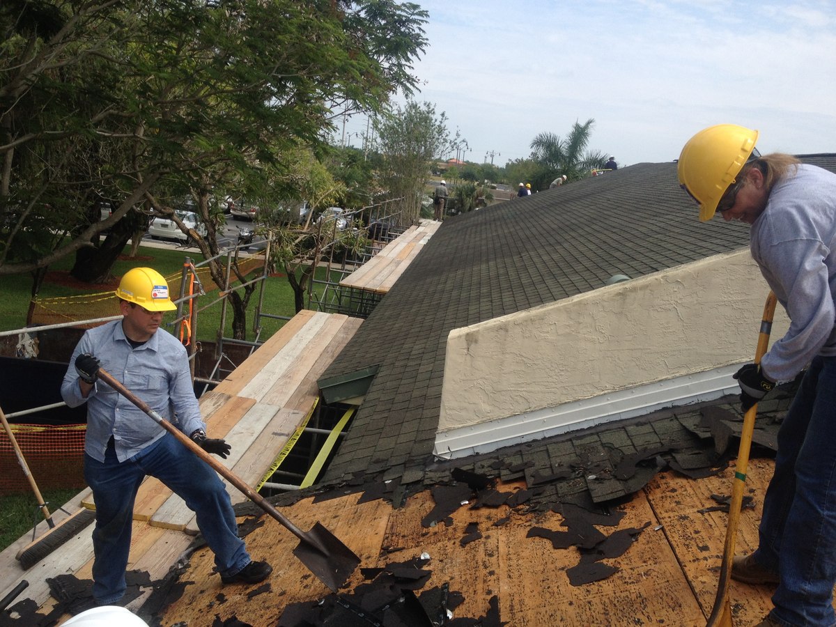Two roofers in yellow hard hats stripping old asphalt shingles with flat shovels from a residential gable roof under blue sky.