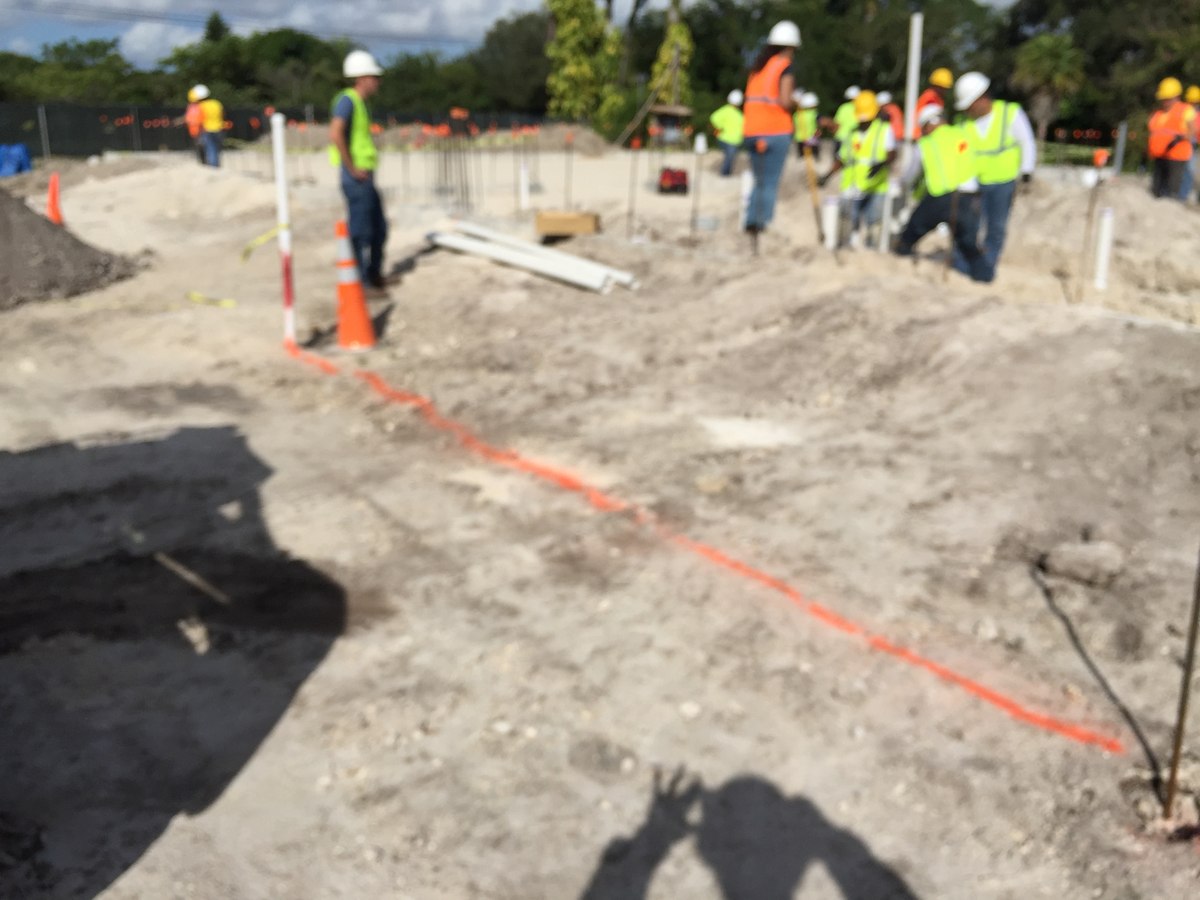 Crew of ten in orange and yellow high-vis vests laying out rebar dowels and PVC conduit on a commercial foundation lot.