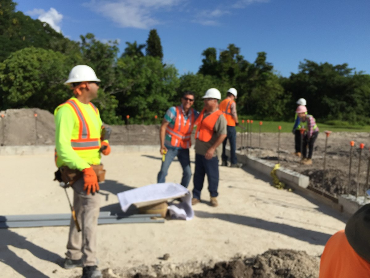 Worker in red hard hat and lime-orange high-vis in foreground with two crew conferring over plans on a commercial foundation slab.