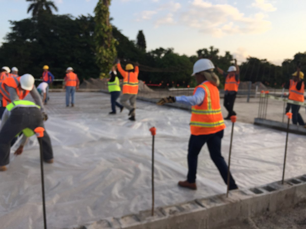Slab pour-day at sunset with vapor barrier and rebar dowels, nine crew in high-vis vests directing the pour on a commercial foundation.