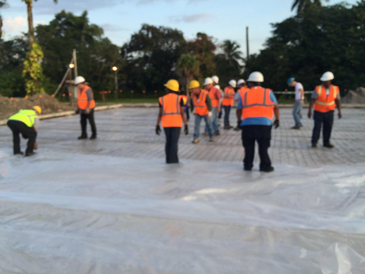 Crew of ten in orange and yellow high-vis vests and hard hats laying out a slab pour over vapor barrier on a commercial site at golden hour.