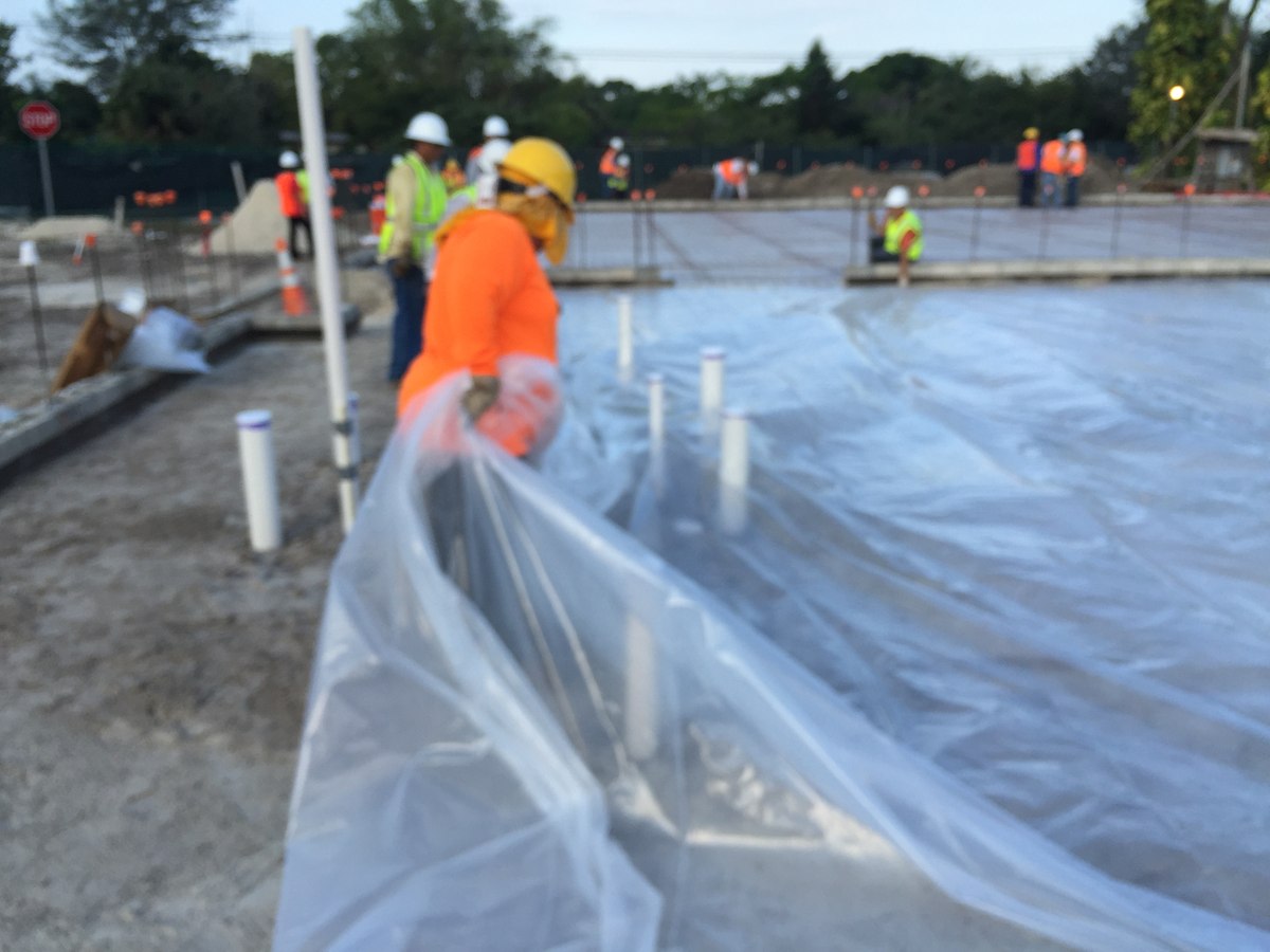 Worker in orange sun-shirt and yellow hard hat pulling a vapor-barrier sheet across a slab while crew assists in background.