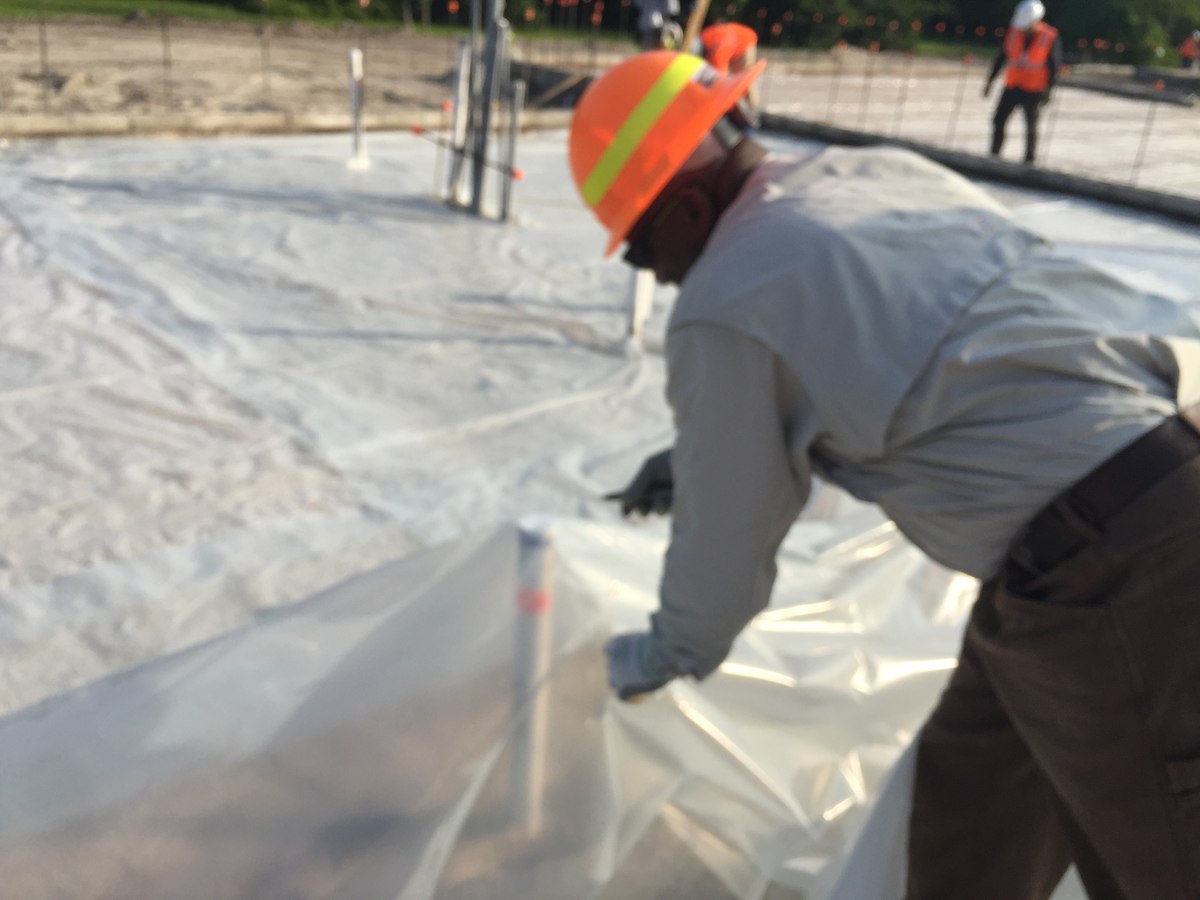 Worker in orange hard hat and gray long-sleeve placing dowel through vapor barrier on a commercial foundation slab.