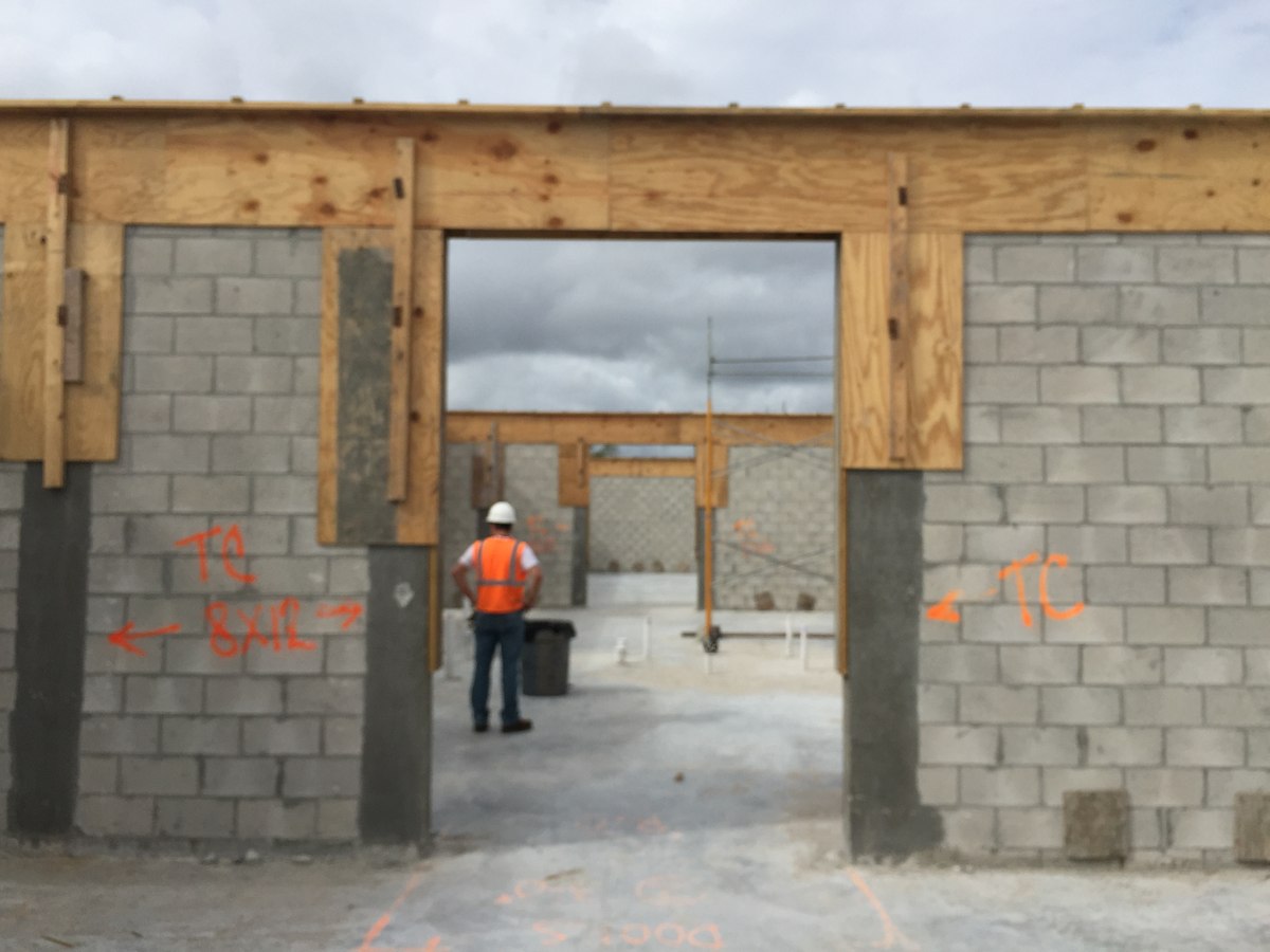 Crew member in orange vest and white hard hat reviewing a CMU-and-plywood structural opening with bond-beam markings.