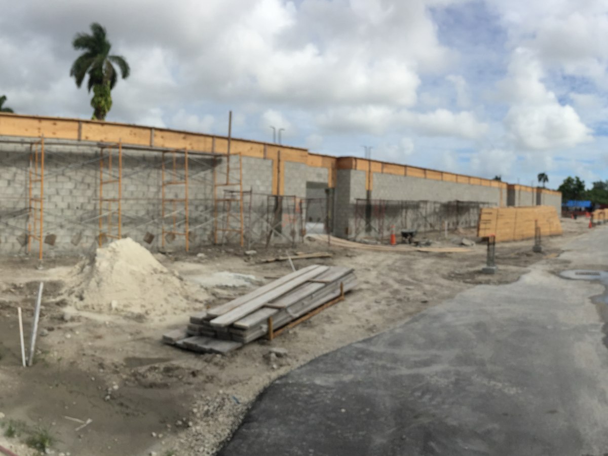 Wide panoramic view of a large commercial CMU shell with scaffolding throughout, sand piles, lumber stacks, and porta-johns.