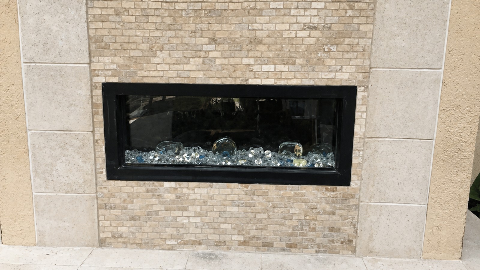Travertine-and-running-bond-stone outdoor fireplace surround with glass fire-media insert, finished outdoor feature.