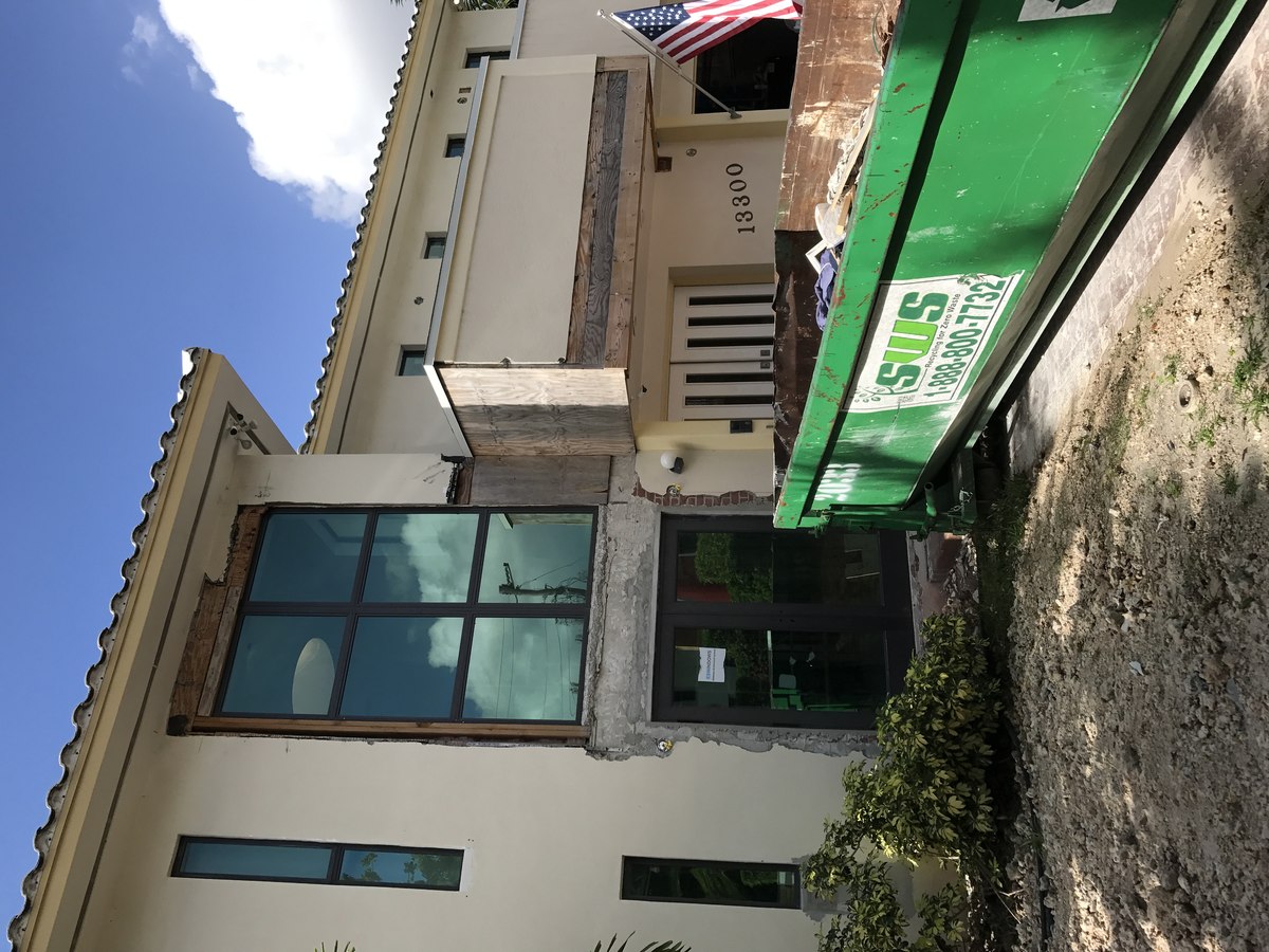 Two-story Mediterranean home mid-restoration — entry column stripped to concrete and lath with green dumpster on the lawn and US flag flying.