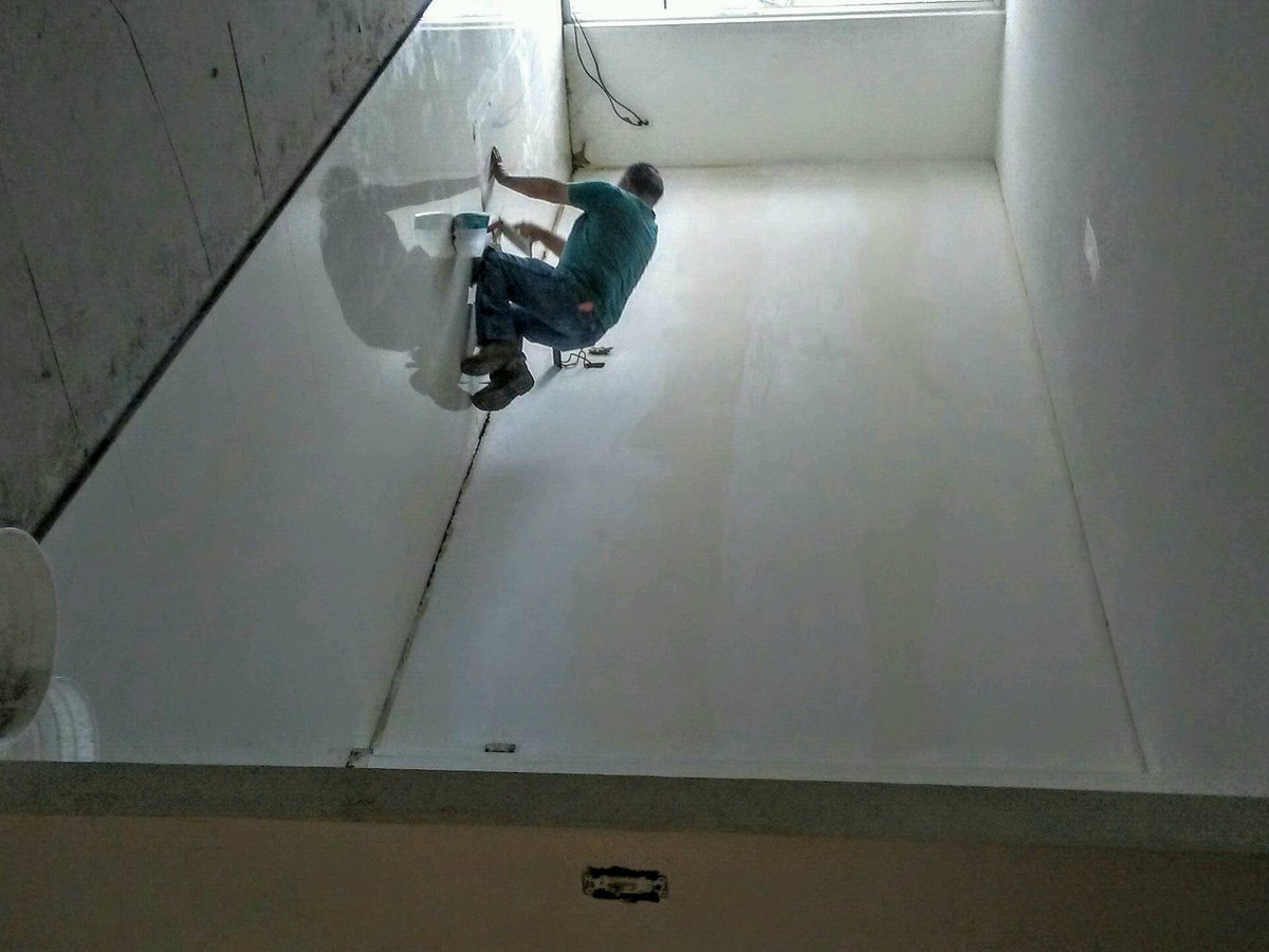 JEGC-branded crew member smooth-coats fresh drywall in a long interior renovation room, light from end window.