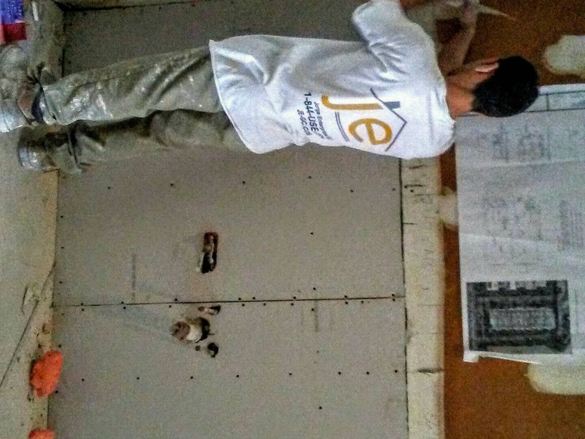 Crew member in white je-gc branded uniform installs drywall panels next to plans pinned to wall — operational scale shot.