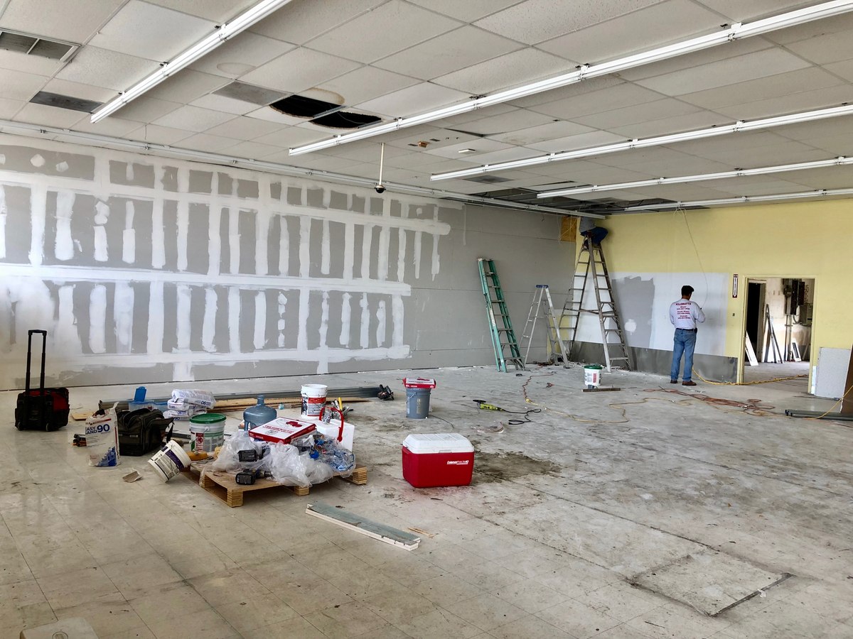 Commercial buildout in progress — taped/mudded drywall, drop-ceiling grid exposed and a worker on a step ladder in a white logo shirt.