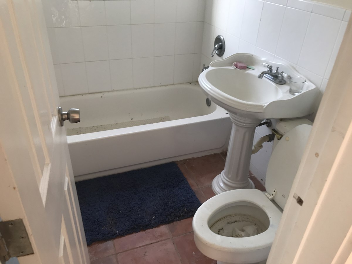 Existing bathroom with old white-tiled walls, stained pedestal sink, and worn terra-cotta tile floor — before our renovation.