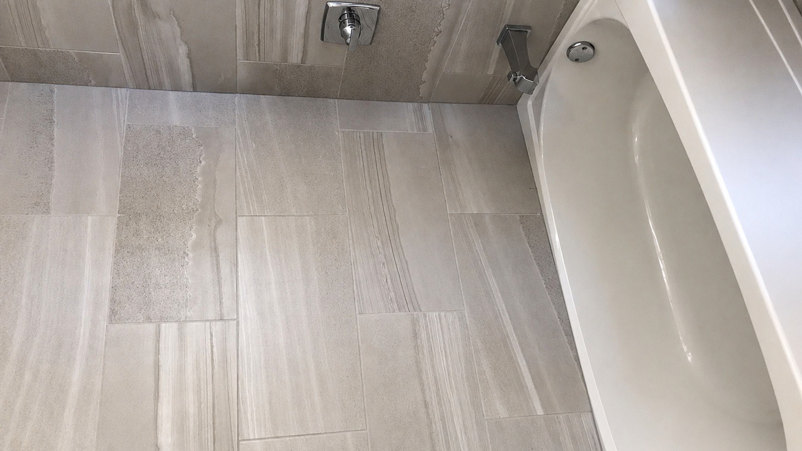 Large-format porcelain tile floor and shower wall meet a deep soaker tub; chrome shower control trim and grab fitting visible.