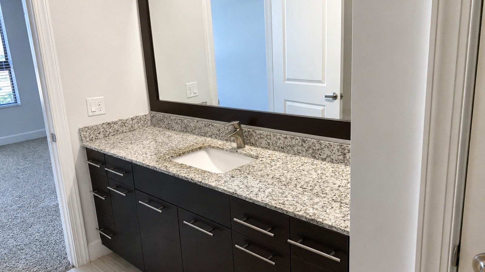 Long espresso vanity with white-and-grey granite top and dark wood-framed mirror, plank tile floor, view through to carpeted bedroom beyond.