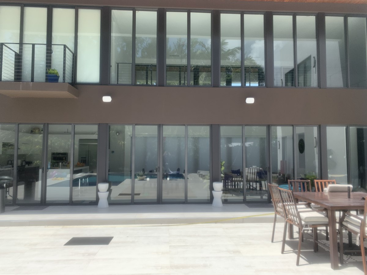 Modern two-story luxury residence rear elevation: floor-to-ceiling glass walls, brown stucco, infinity pool through the glass, travertine patio with dining set.