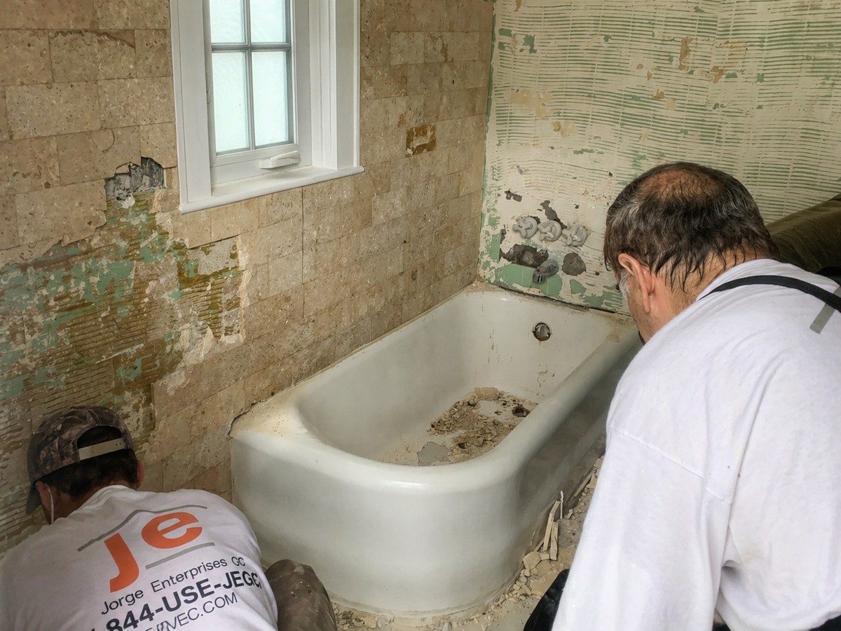 Two JE-GC workers in branded shirts demolishing dated tile around a cast-iron bathtub, exposed wall adhesive and rubble across the floor.