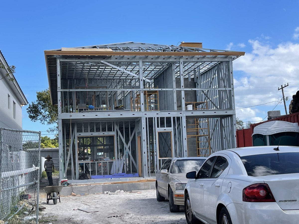 Two-story light-gauge steel structural framing for a new build, with a worker mixing concrete in a wheelbarrow at left.