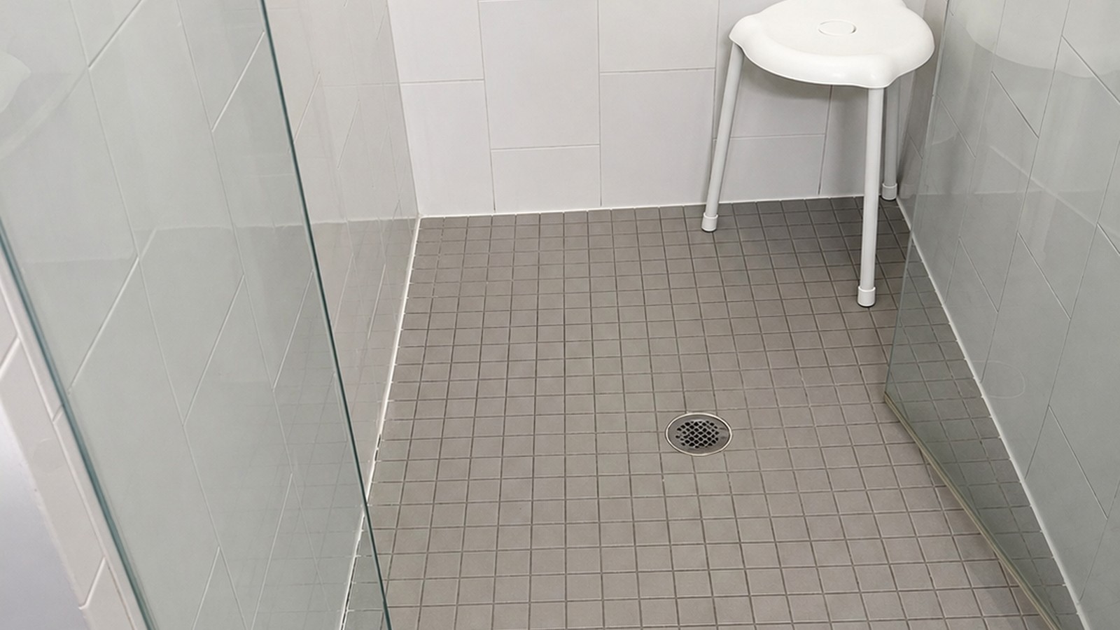 Compact ADA shower with white square tile, frameless glass partition, marble curb, brushed-nickel grab bar, and accessibility seat.