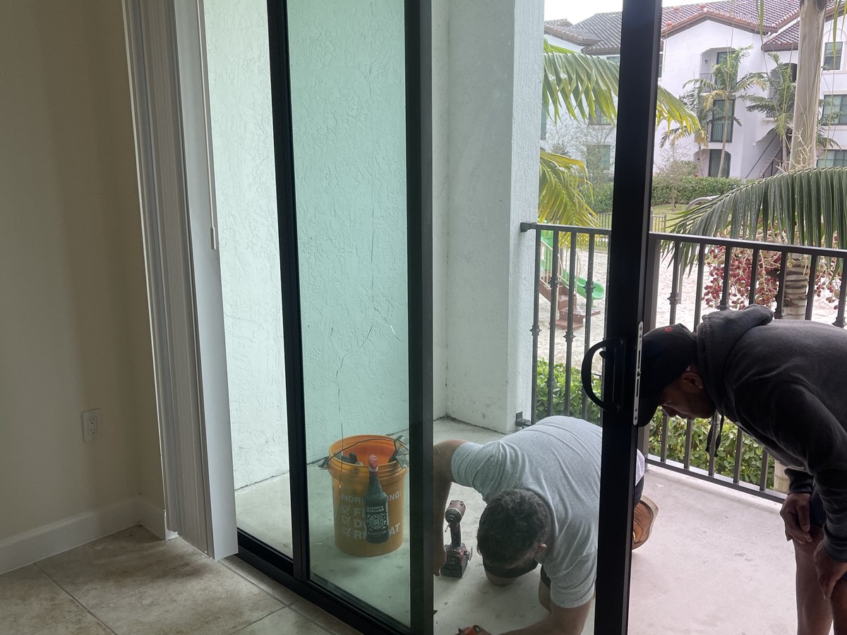 Two-person crew aligns and seals the bottom track of a tall impact slider on a condo balcony, courtyard view beyond.