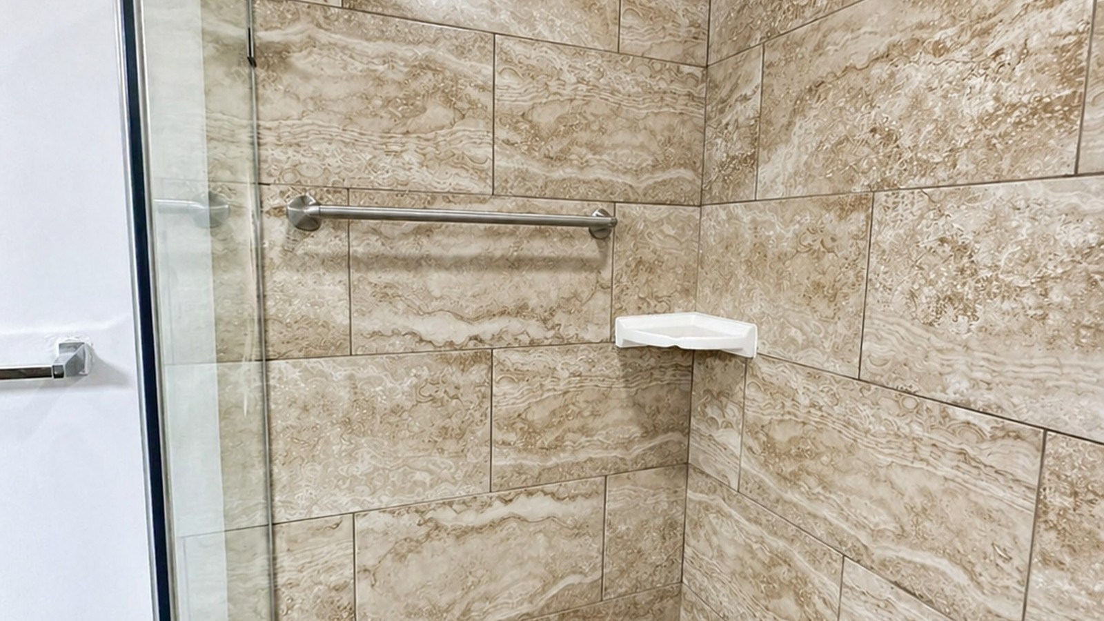 Travertine-look porcelain tile shower with brushed-nickel grab bar and corner shelf, mosaic floor visible.