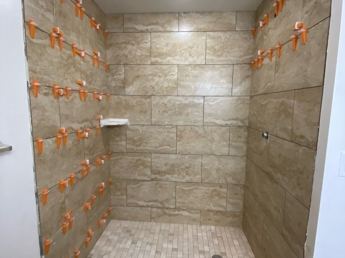 Travertine-look porcelain tile installed with orange leveling clips on shower walls, mosaic floor laid, ready for grout.