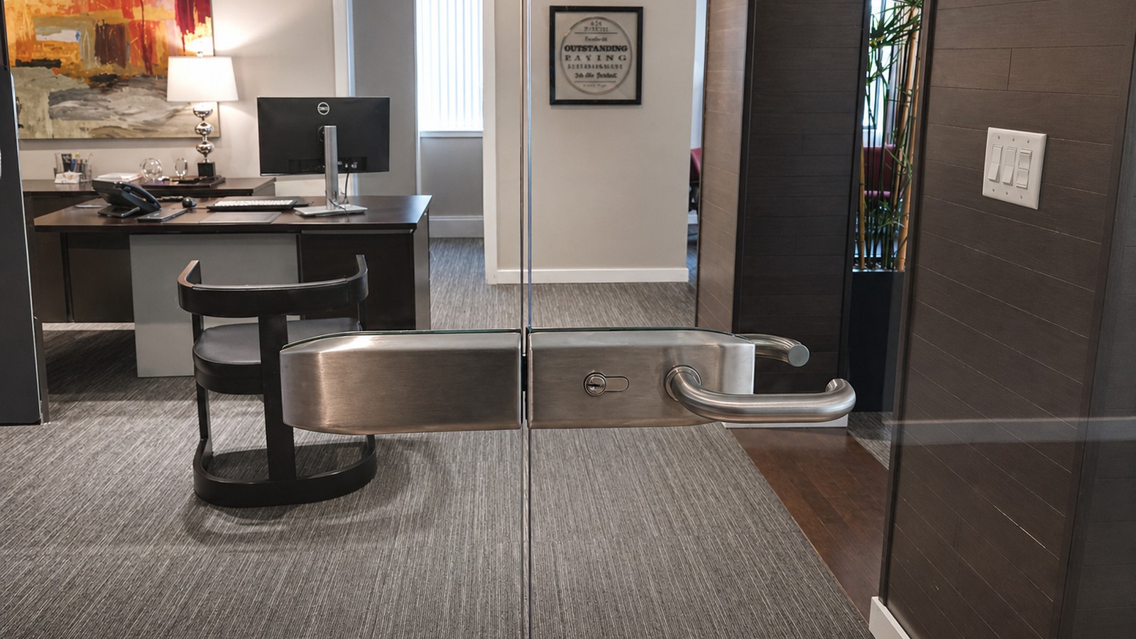 Frameless glass partition with stainless push-pull hardware separates a finished office area with art and dark plank floor.